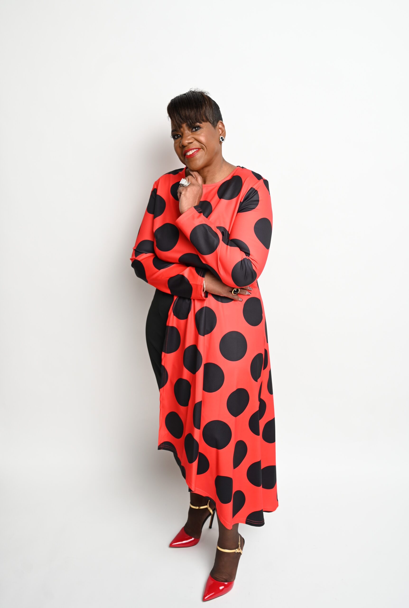 A photograph of Janet Tonkins in a long flowing red top with big black polka dots in front of a bright white background.